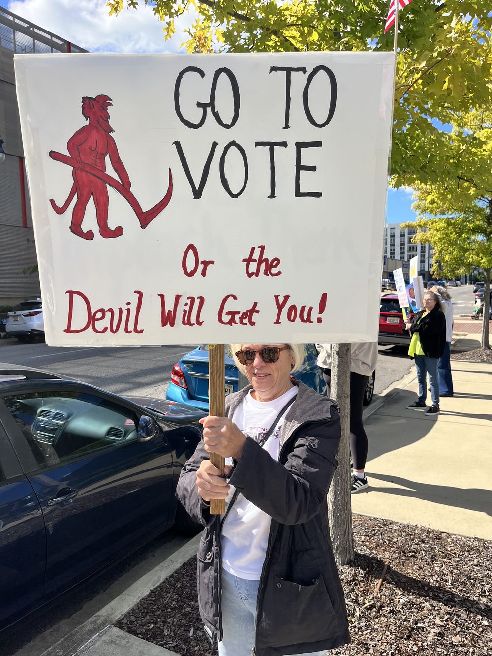 A woman holding a sign that reads Go to Vote or the Devil Will Get You.