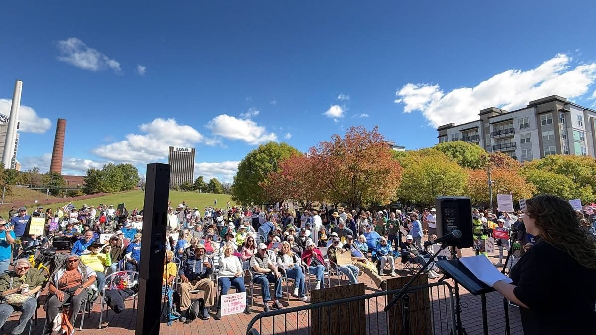 A crowd seated on the lawn at Railroad Park in Birmingham, facing a stage, under a blue sky with autumn trees.