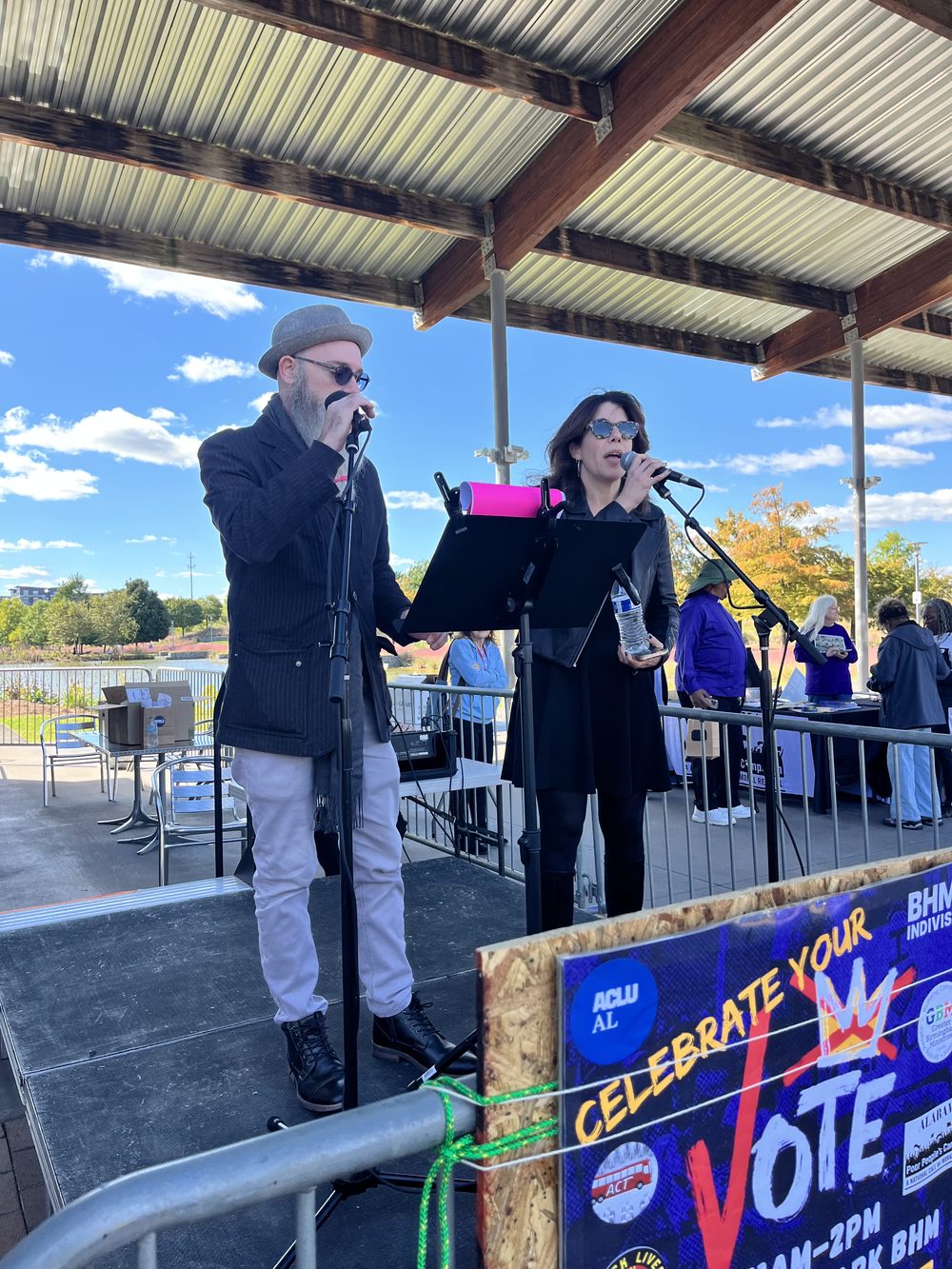 Two people speaking at microphones on the Railroad Park stage, with a Celebrate Your Vote banner in the foreground.