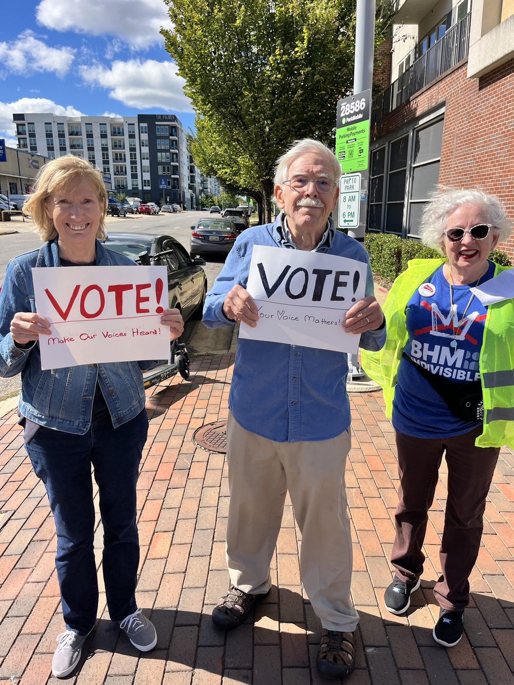 Three people on a brick sidewalk holding hand-lettered VOTE signs.